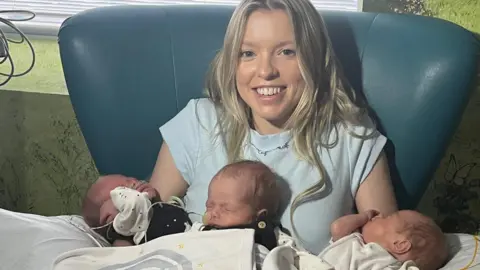 Shannon Page smiling and wearing a white top with Cruz, Aljo and Enzo on her lap. The boys are in matching black dungarees with white blankets over them and some tubes