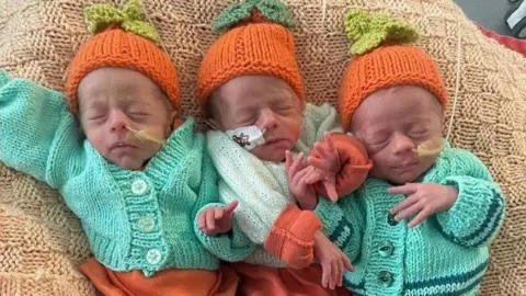 Ace & Shannon Page The trio of triplets lie together on a beige-coloured blanket. They are wearing matching green cardigans and knitted orange hats. All three are lying on their backs and have their eyes closed