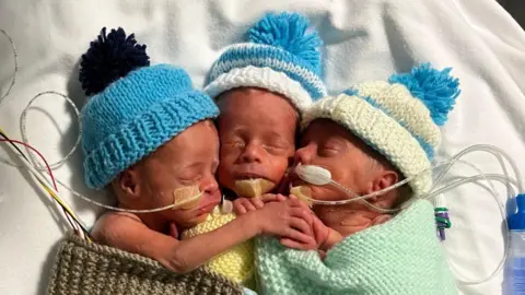 Ace & Shannon Page The trio lying next to each other holding hands in the hospital unit. They are all wearing blue hats and have medical tubes attached, with one baby wrapped in a brown blanket, one in yellow and one in green